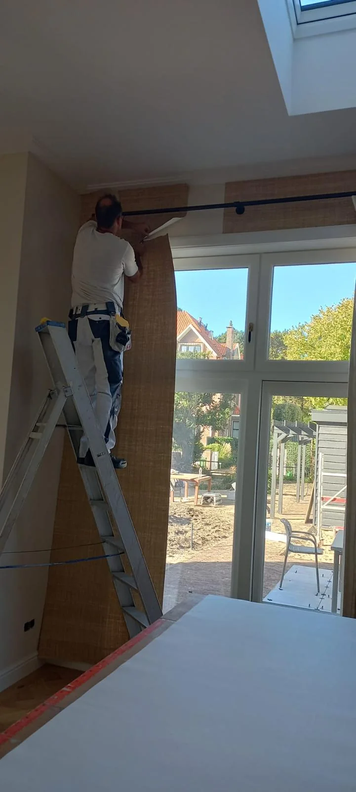 Een behangbaan wordt nauwkeurig bij een grote raampartij aangebracht.